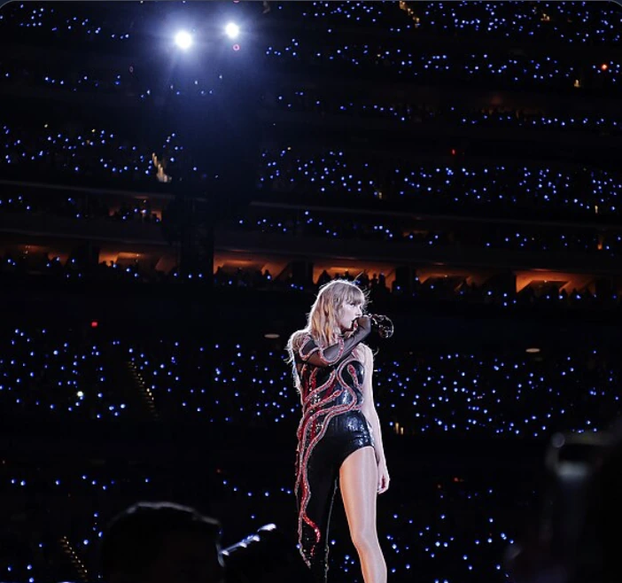 Taylor Swift performing alone on a brightly lit stadium stage before a large audience holding blue lights